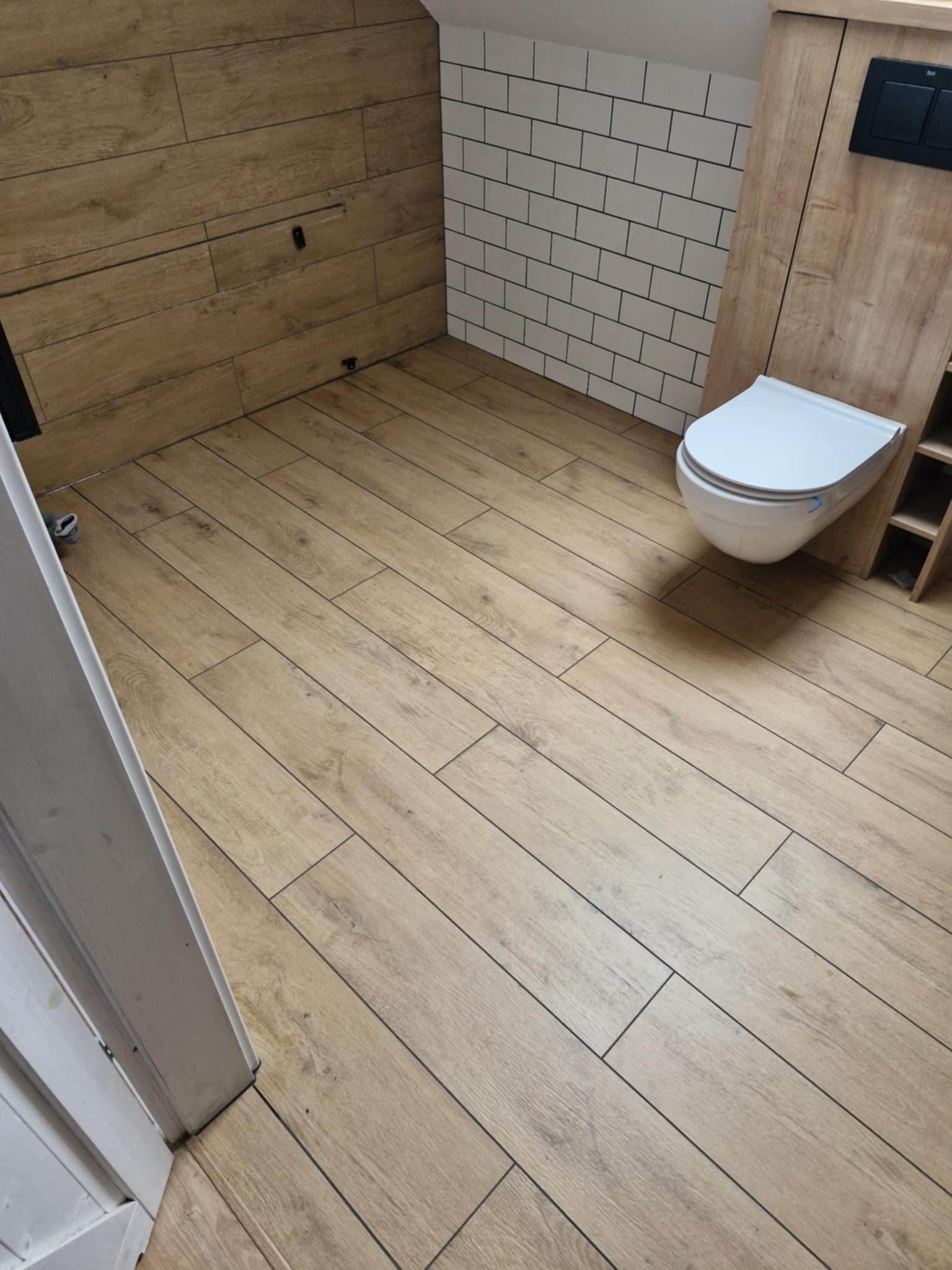 Wykończona łazienka z drewnopodobną podłogą i ścianą z płytek imitujących cegłę. Podwieszana toaleta z zabudową. Ukośne ściany dodają charakteru.