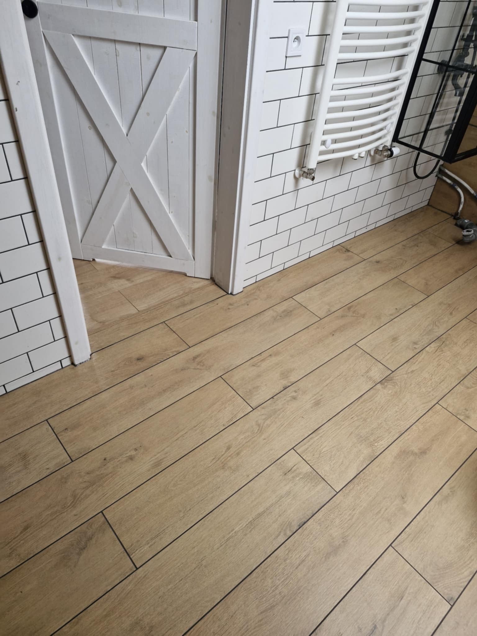 Jasne panele podłogowe w łazience z białą, drewnianą, przesuwną  drzwi i grzejnikiem na ścianie wyłożonej białymi płytkami imitującymi cegłę.