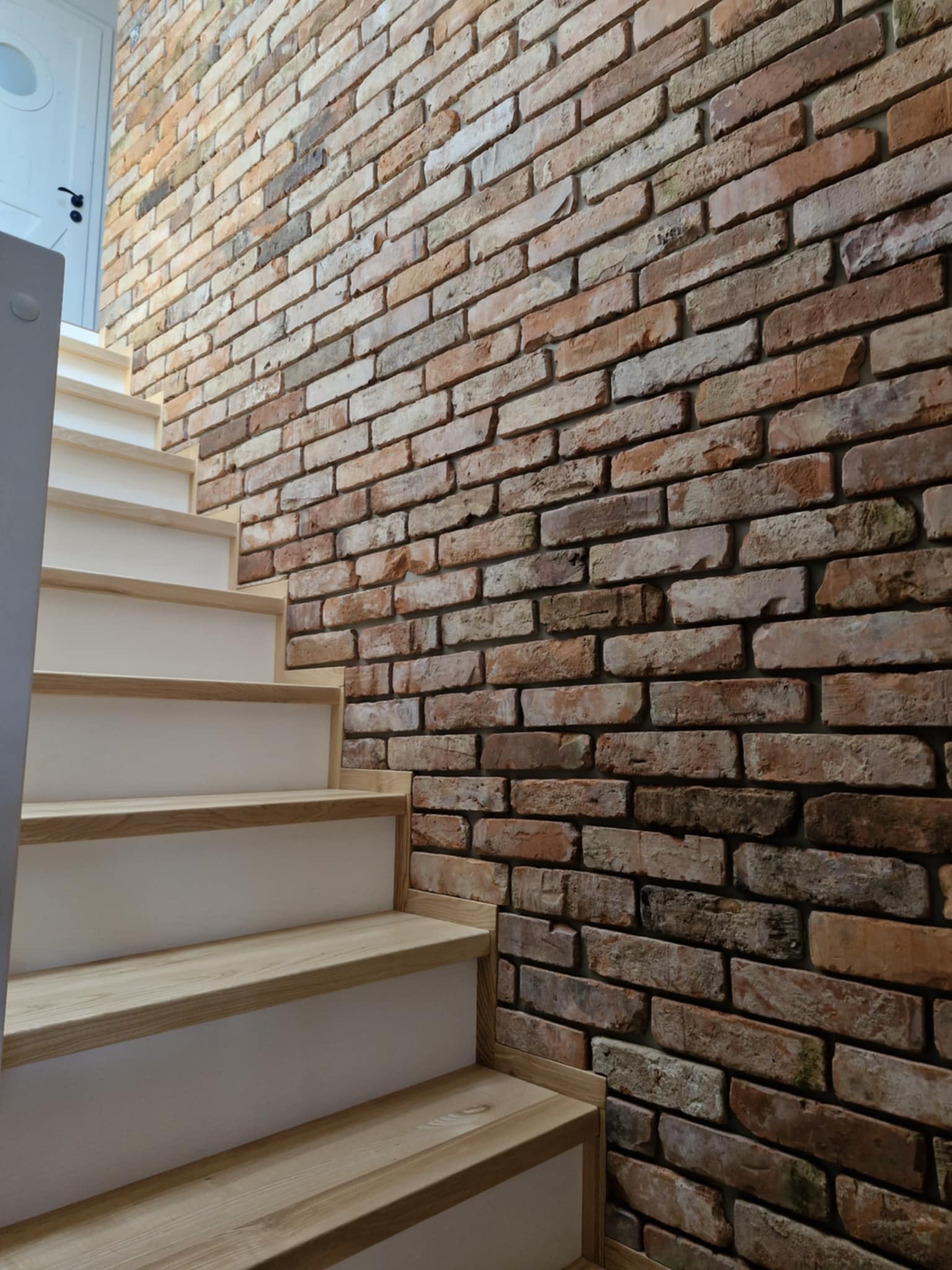 Schody z drewnianymi stopniami i białą podstopnicą, przylegające do ściany z dekoracyjnej cegły w odcieniach brązu i beżu, oświetlone naturalnym światłem.