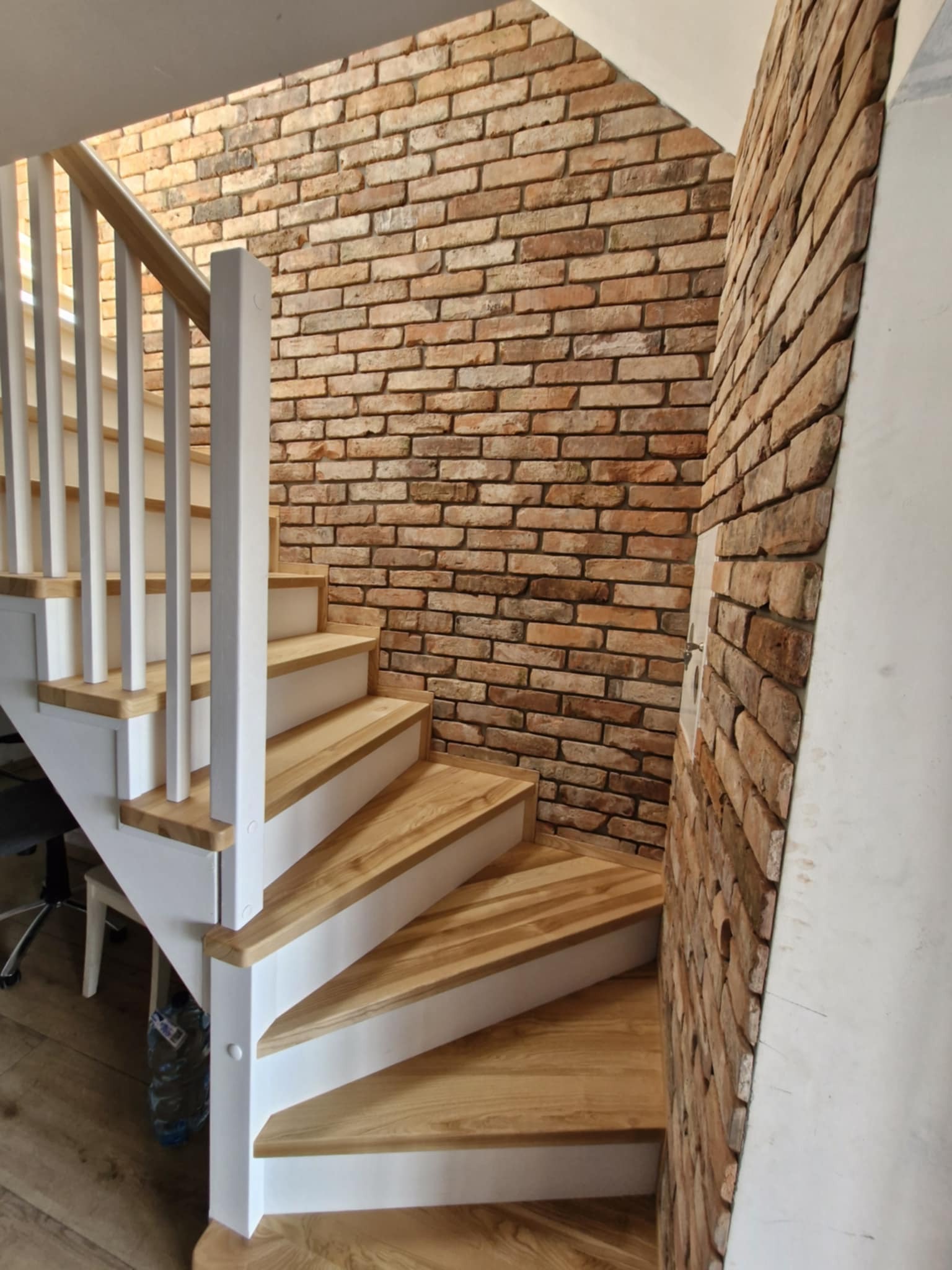 Drewniane schody z białymi balustradami, obok ściana z cegły w odcieniach brązu i beżu. Widok z dołu, ukazujący zakręt schodów i fakturę ceglanej ściany.