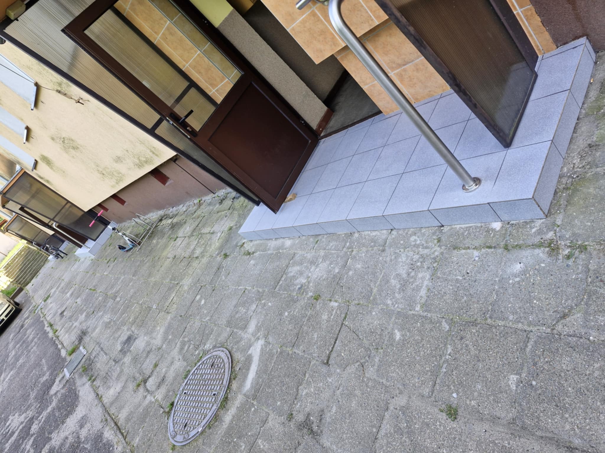 Wejście do budynku z brązowymi drzwiami i szarymi płytkami na schodach oraz chodniku. Widoczna metalowa poręcz i fragment elewacji. Ujęcie z dołu, obrócone.