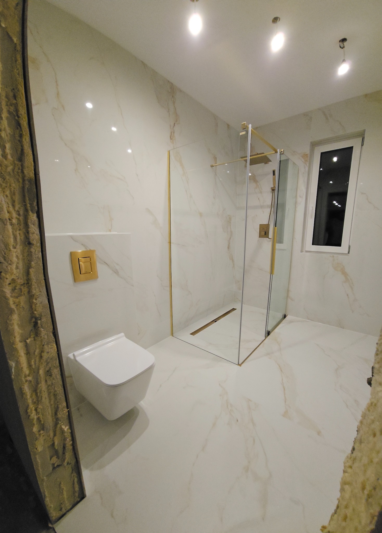 Elegancka łazienka z kabiną prysznicową ze złotymi detalami, podwieszaną toaletą i marmurowymi płytkami. Nowoczesny design z oknem.