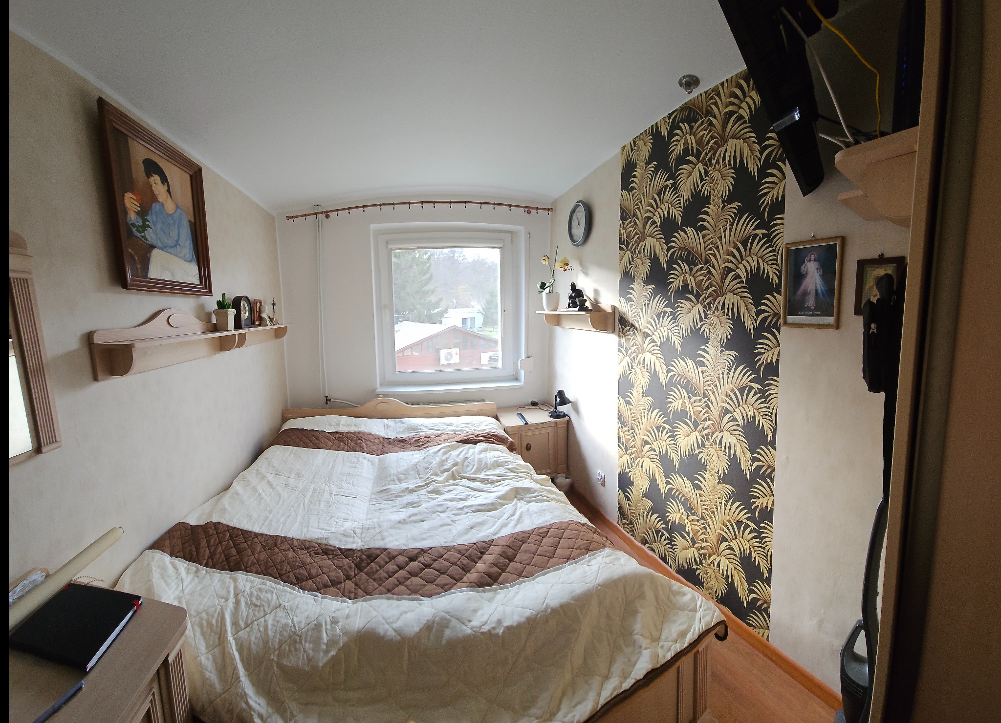 Przytulna sypialnia z łóżkiem, wzorzystą tapetą w palmy i drewnianymi meblami. Na ścianach obrazy i półki z dekoracjami. Widok z okna na zewnątrz.