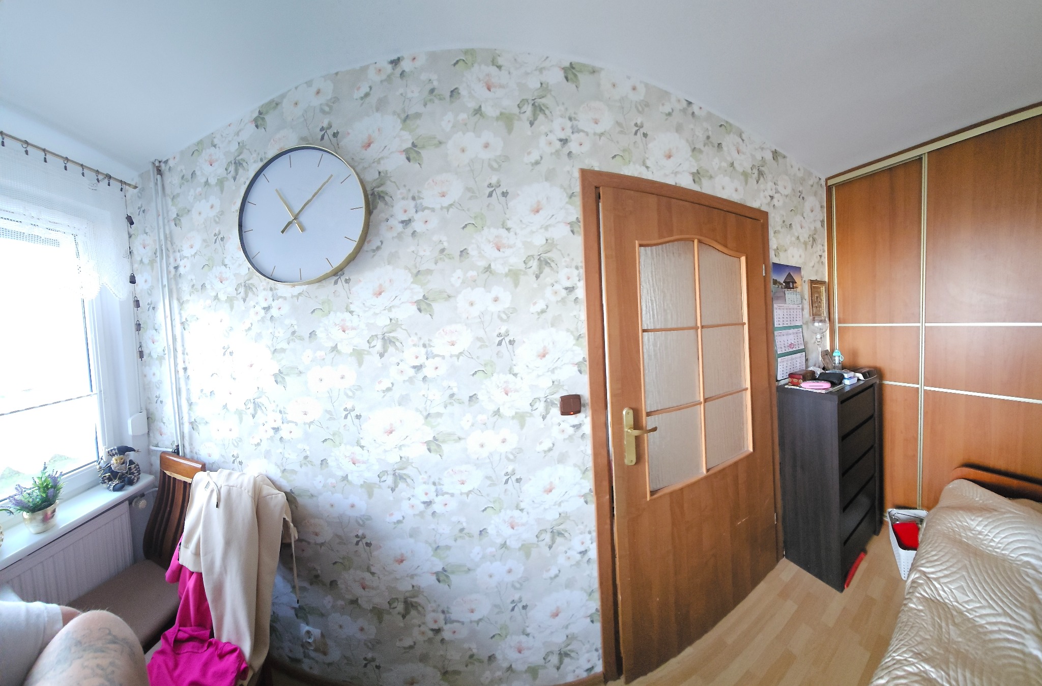 Pokój po remoncie: ściana w kwiaty, drewniane drzwi z szybą, duży zegar na ścianie, szafa przesuwna i komoda. Widok na okno z parapetem i kwiatami.