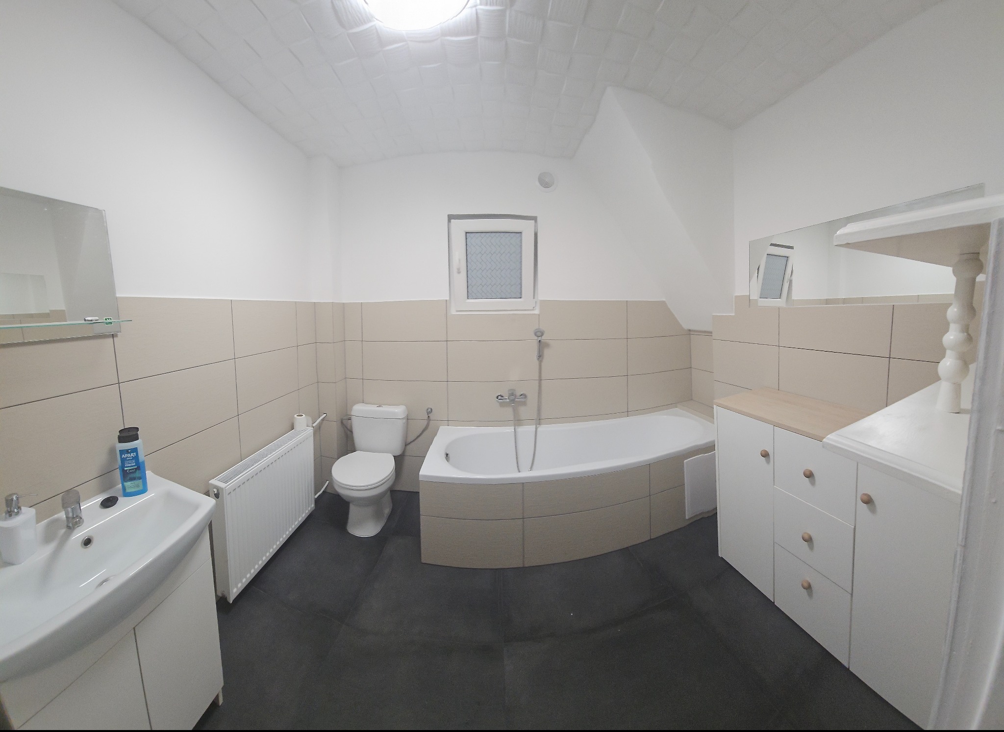 Jasna łazienka z beżowymi płytkami, białą armaturą i ciemną podłogą. Umywalka z szafką, toaleta, wanna obudowana płytkami i biała szafka z drewnianym blatem.