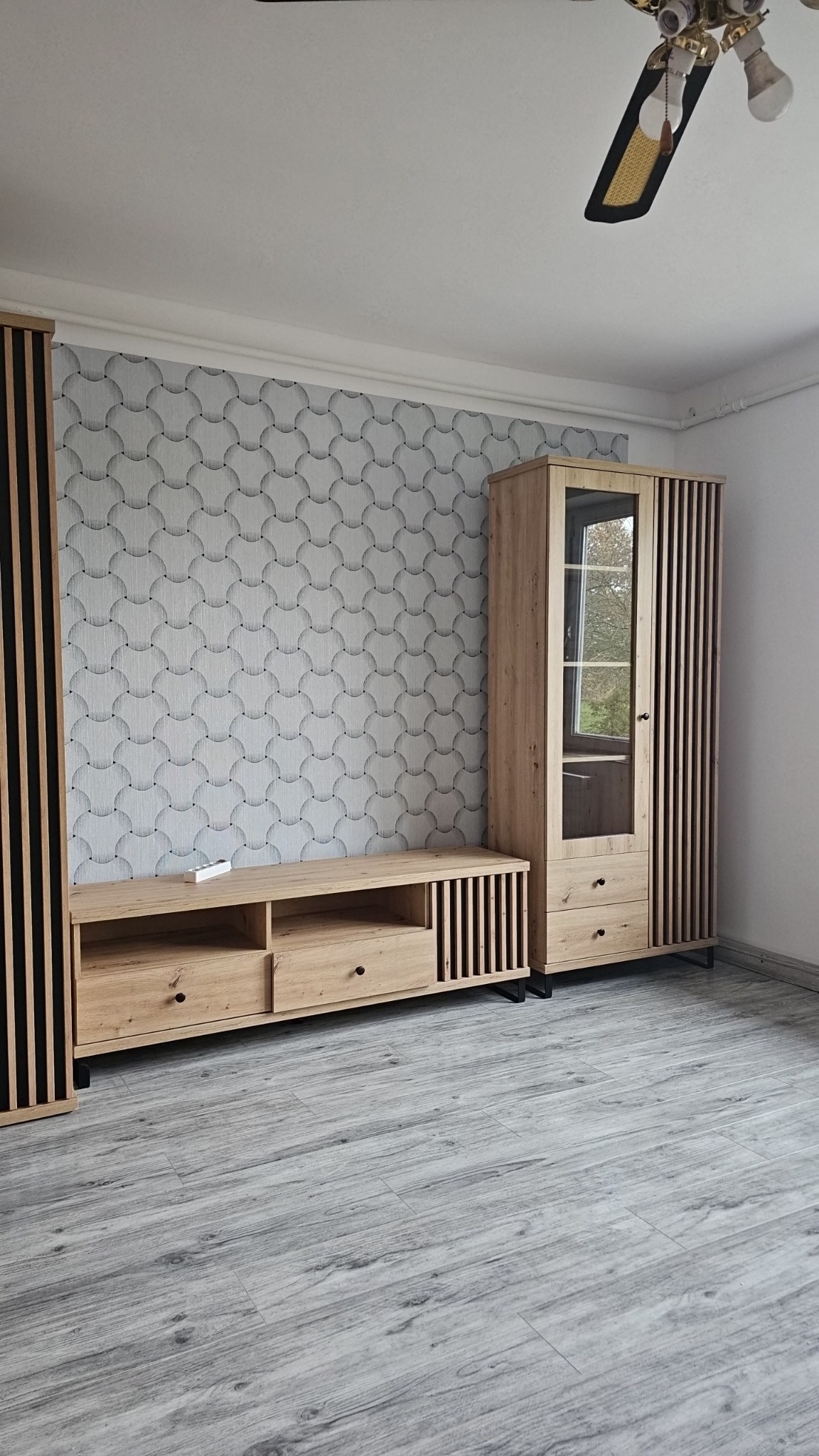 Jasne drewniane meble z ryflowanymi elementami na tle wzorzystej tapety i szarych paneli podłogowych. Widoczny fragment sufitu z wentylatorem.