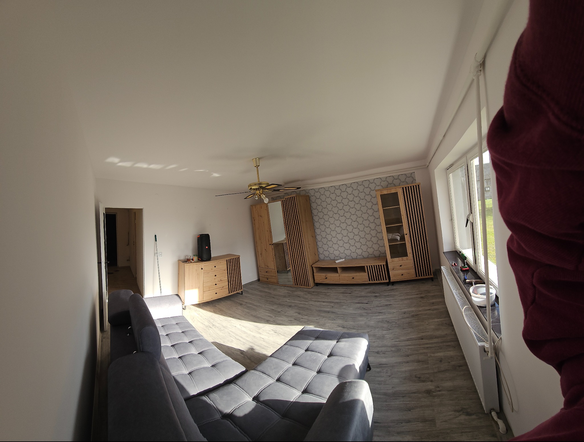 Jasne wnętrze po remoncie: szara sofa, drewniane meble, wzorzysta tapeta, okno z narzędziami. Widok na odnowiony salon z elementami wykończeniowymi.