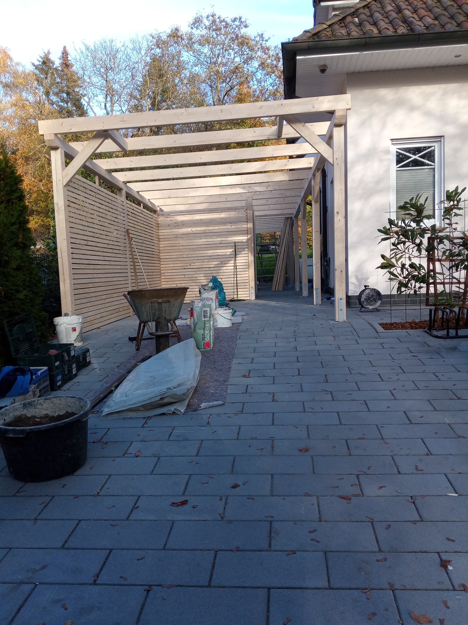 Drewniana konstrukcja z zadaszeniem w trakcie budowy na posesji, widoczne narzędzia i materiały budowlane, kostka brukowa na podłożu.