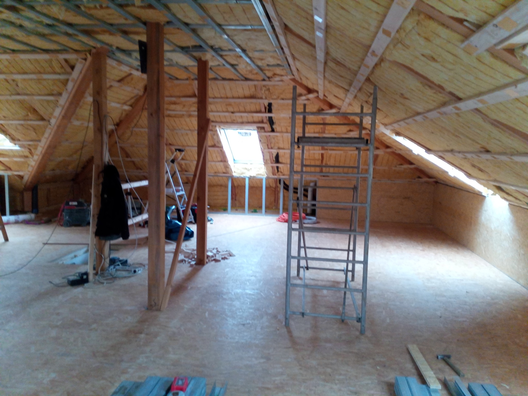 Ocieplone poddasze z oknem dachowym w trakcie adaptacji, widoczne drewniane elementy konstrukcyjne, izolacja, metalowa drabina i narzędzia na podłodze.