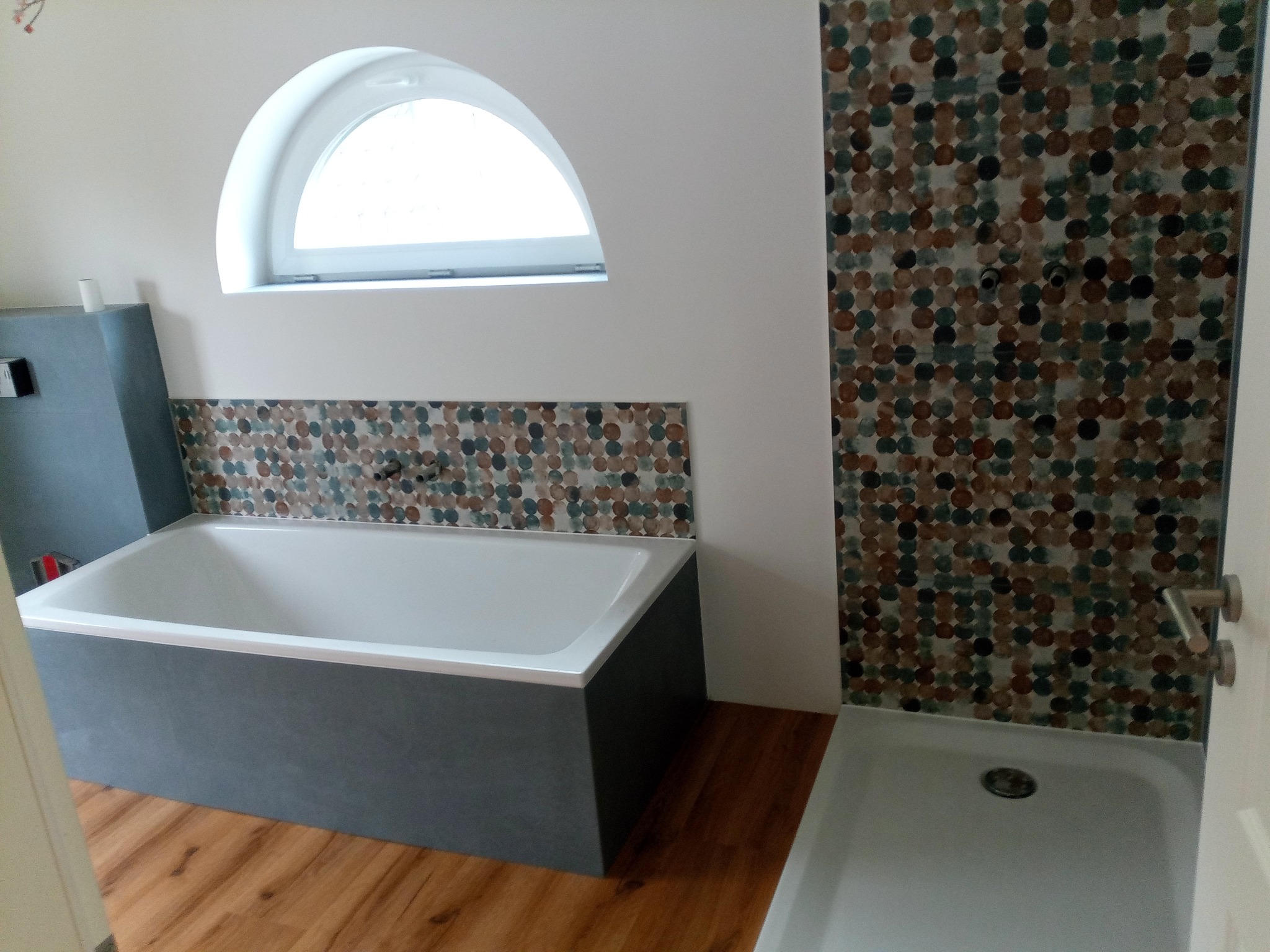 Wykończona łazienka z wanną obudowaną szarą płytą, mozaiką na ścianie i drewnianą podłogą. Okno w kształcie półkola nad wanną. Nowoczesny design.