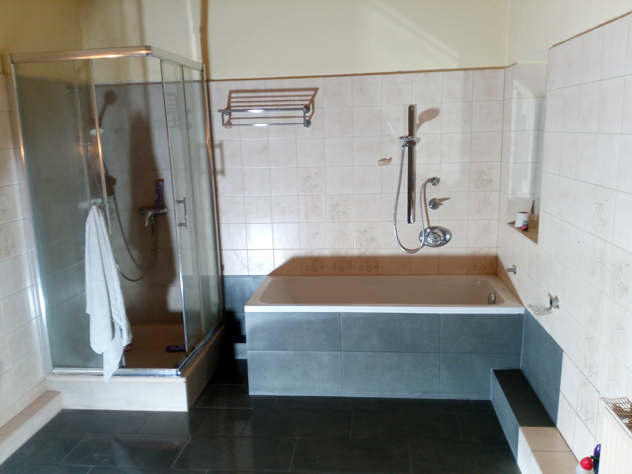 Wyremontowana łazienka z kabiną prysznicową, wanną obudowaną szarymi płytkami i ciemną podłogą. Na ścianach jasne płytki z delikatnym wzorem. Widoczny ręcznik w kabinie.