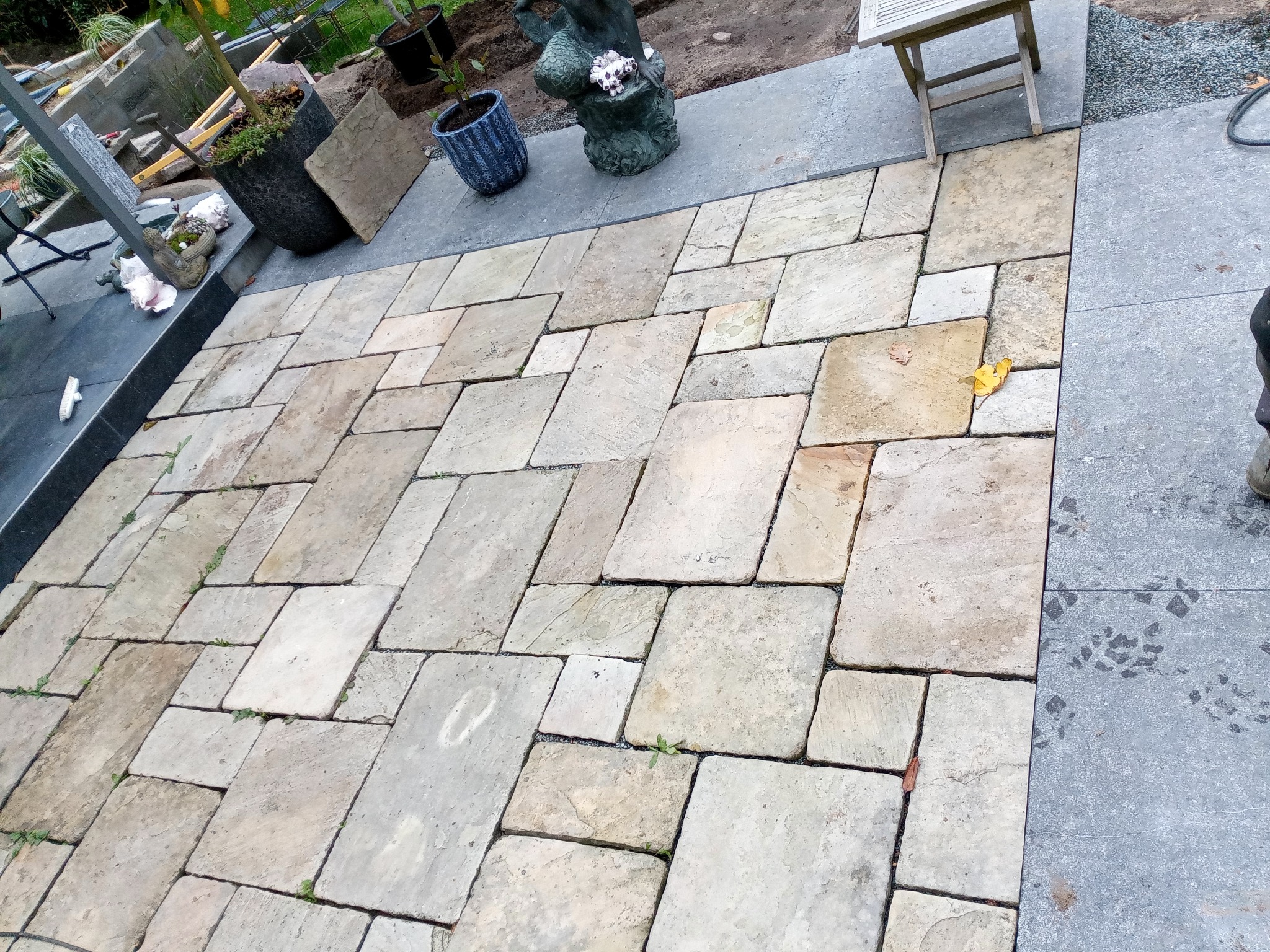 Ułożona, nieregularna nawierzchnia z jasnych, kwadratowych płyt kamiennych na zewnątrz, z elementami dekoracyjnymi i roślinnością w tle. Widoczne ślady użytkowania.