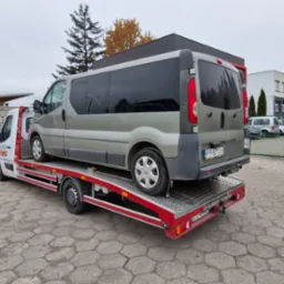 Srebrny van na czerwono-białej platformie transportowej, widok z tyłu, na tle budynków i drzew, z widocznym logo firmy pomocy drogowej na kabinie.