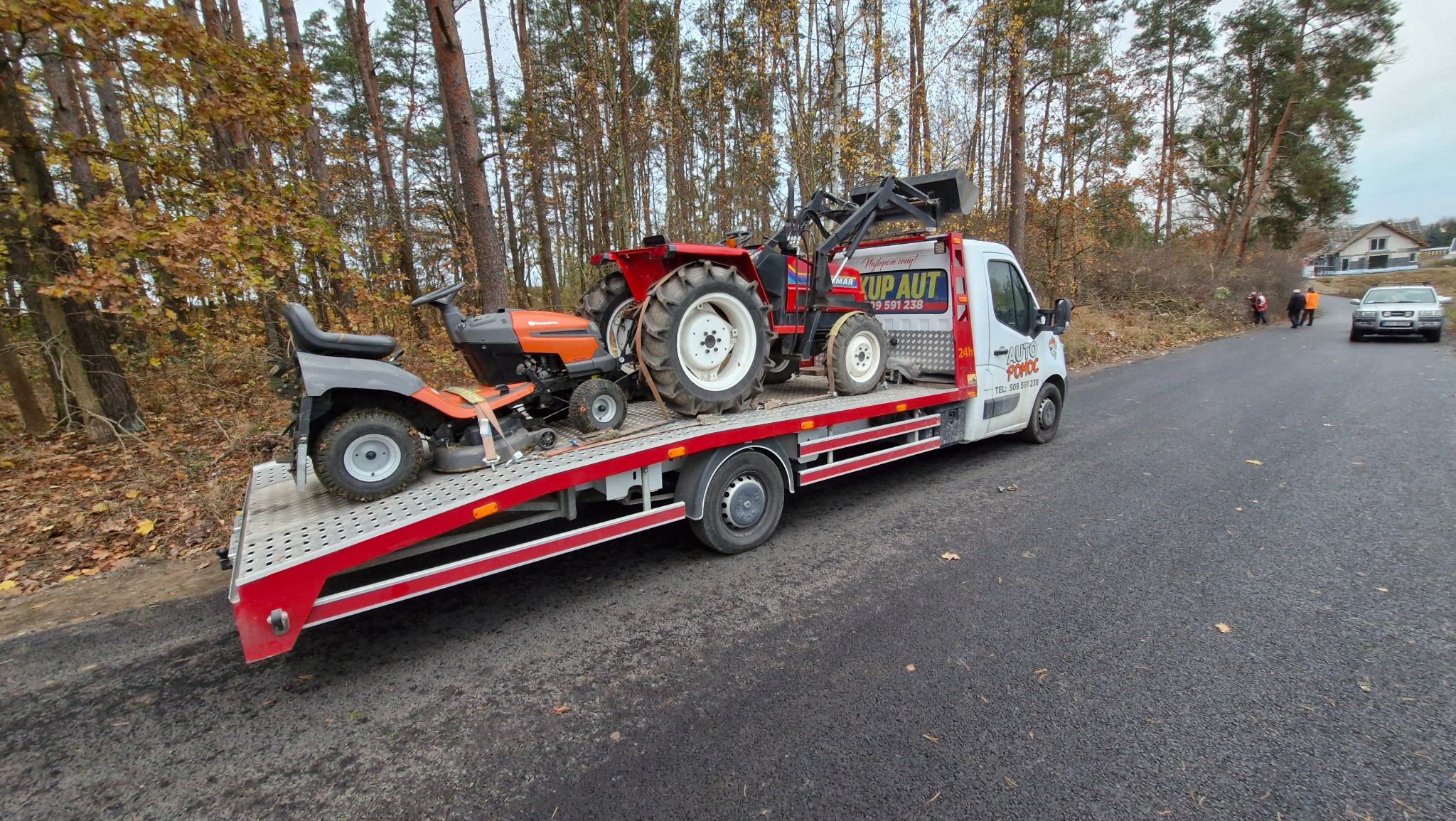 Transport małego traktora rolniczego i kosiarki do trawy na platformie pojazdu pomocy drogowej z widocznym logo firmy na zabudowie, las w tle.