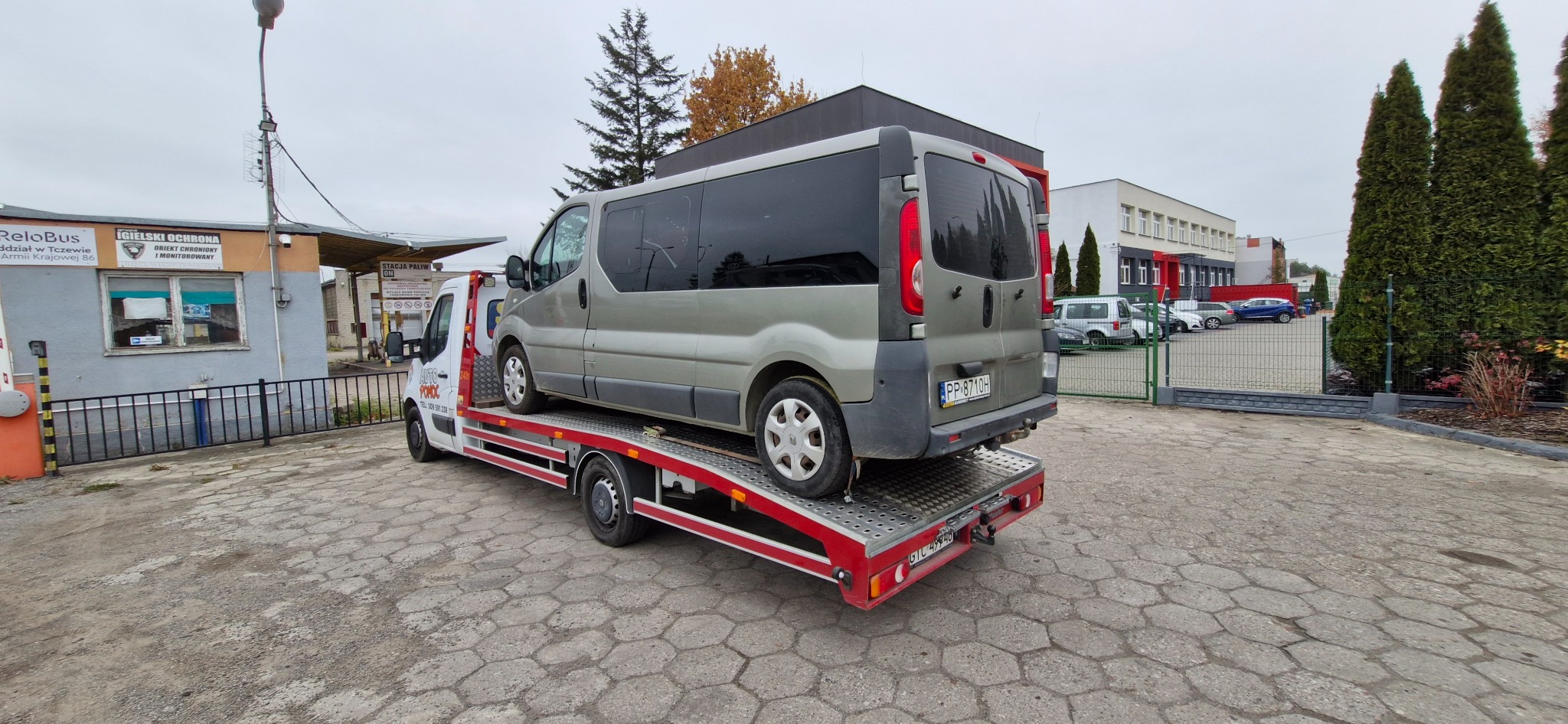 Srebrny van na czerwono-białej platformie transportowej, widok z tyłu, na tle budynków i drzew, z widocznym logo firmy pomocy drogowej na kabinie.