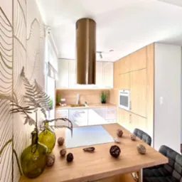 Nowoczesna kuchnia z drewnianą wyspą i okrągłym, miedzianym okapem, zielone wazony z paprocią na tle ściany z motywem liści.