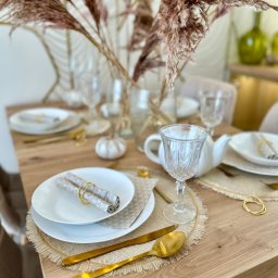 PROJEKTYWNA Agnieszka Michniewicz-Kupś - Uroczysty stół w Koluszkach, nakryty białymi talerzami z serwetkami w złotych pierścieniach, złotymi sztućcami, szklankami na nóżce oraz dekoracją z suszonych traw w wazonie, tworząc elegancką...