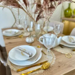 Uroczysty stół w Koluszkach, nakryty białymi talerzami z serwetkami w złotych pierścieniach, złotymi sztućcami, szklankami na nóżce oraz dekoracją z suszonych traw w wazonie, tworząc elegancką...