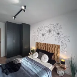 Nowoczesna sypialnia z ciemnymi meblami, drewnianym parkietem w jodełkę, łóżkiem z wezgłowiem w kolorze miodowym i kwiatowym motywem graficznym na ścianie.