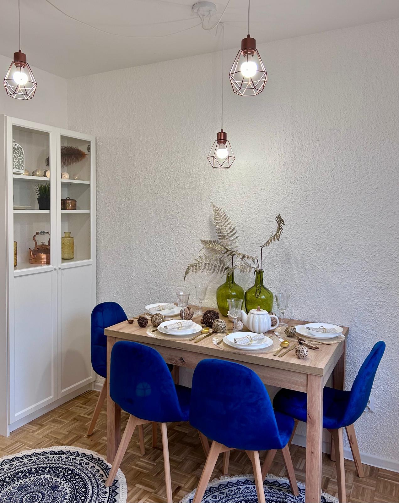 Jadalnia z drewnianym stołem, nakrytym białymi talerzami i złotymi sztućcami, otoczona czterema krzesłami obitymi granatowym welurem. Dekoracje stołu stanowią zielone butelki z suszonymi gałązkami...