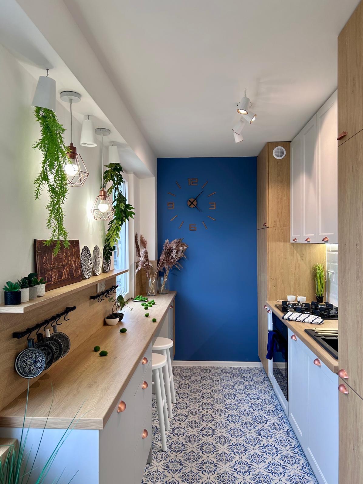 Wąska kuchnia z niebieską ścianą, drewnianym blatem, białymi szafkami z miedzianymi uchwytami, dekoracyjną podłogą w stylu portugalskim, wiszącymi lampami z roślinami i zegarem ściennym.