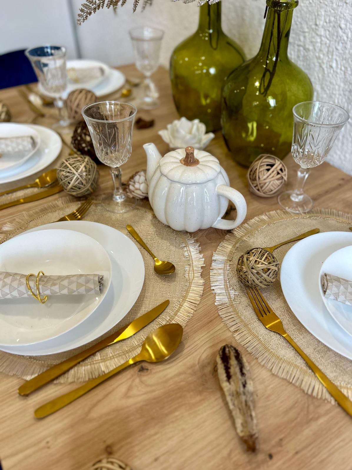 Elegancko nakryty stół z białymi talerzami, złotymi sztućcami, lnianymi podkładkami, szklankami z wzorem, ceramicznym czajnikiem w kształcie dyni i dekoracyjnymi kulami z naturalnych materiałów...