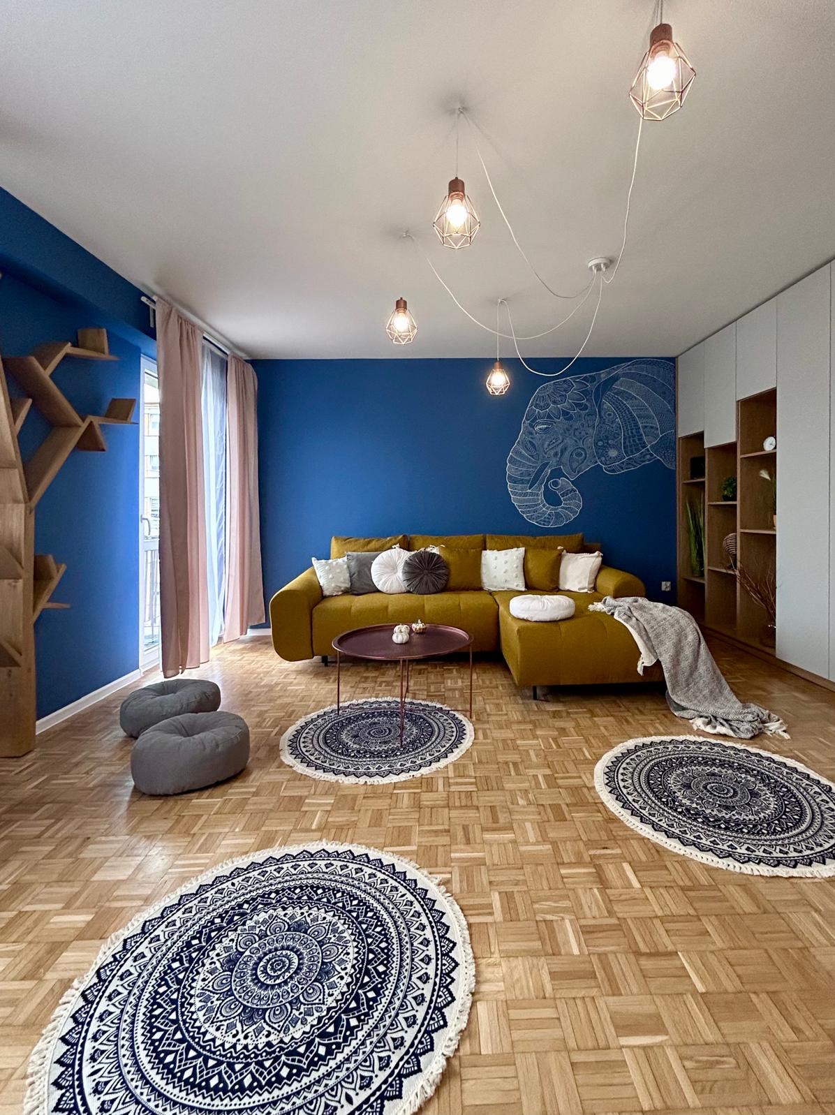 Salon z niebieską ścianą i białym rysunkiem słonia, musztardową sofą, okrągłymi dywanami z mandalami i drewnianą półką w kształcie drzewa.