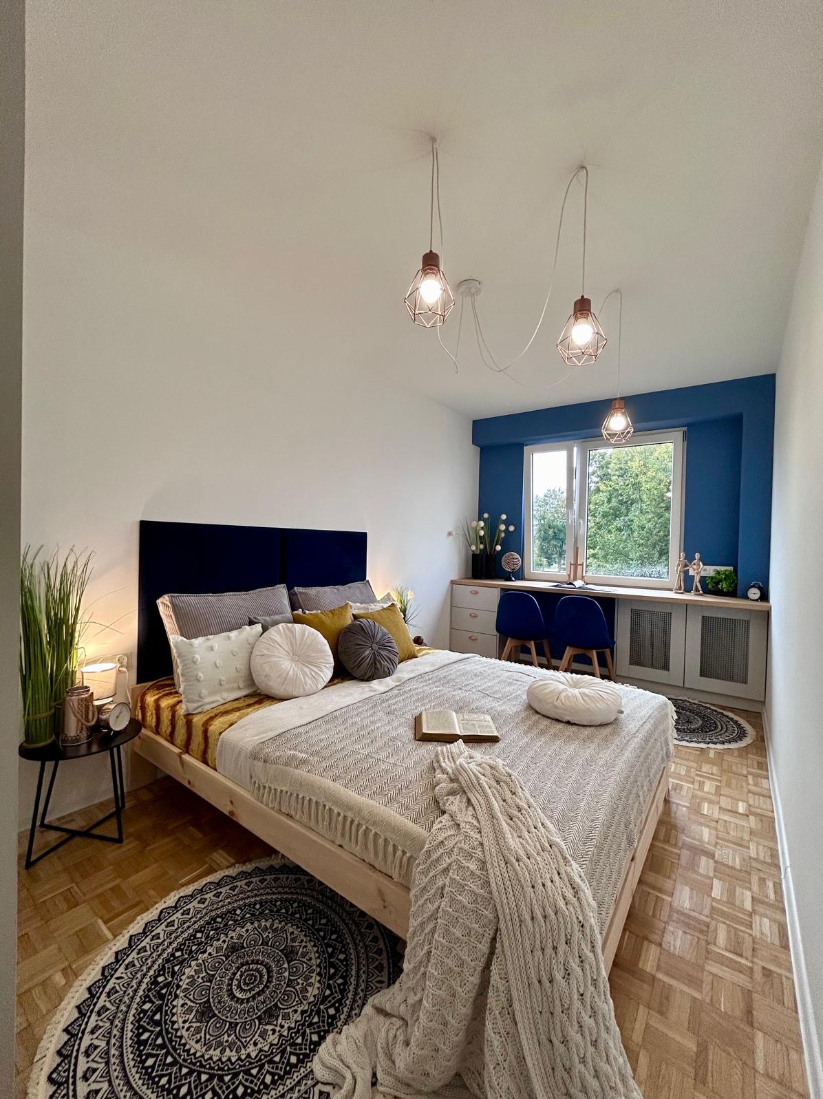 Jasna sypialnia z drewnianym łóżkiem, dekoracyjnymi poduszkami, książką na pościeli i miejscem do pracy z dwoma krzesłami pod oknem, oświetlona trzema lampami drucianymi.