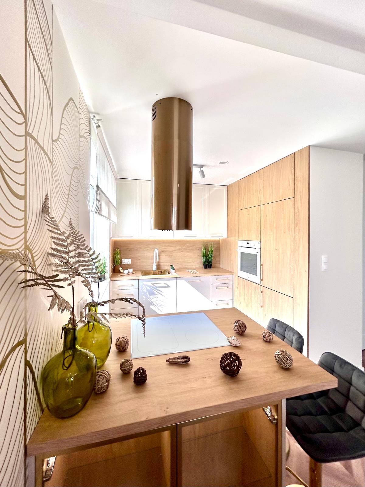 Nowoczesna kuchnia z drewnianą wyspą i okrągłym, miedzianym okapem, zielone wazony z paprocią na tle ściany z motywem liści.