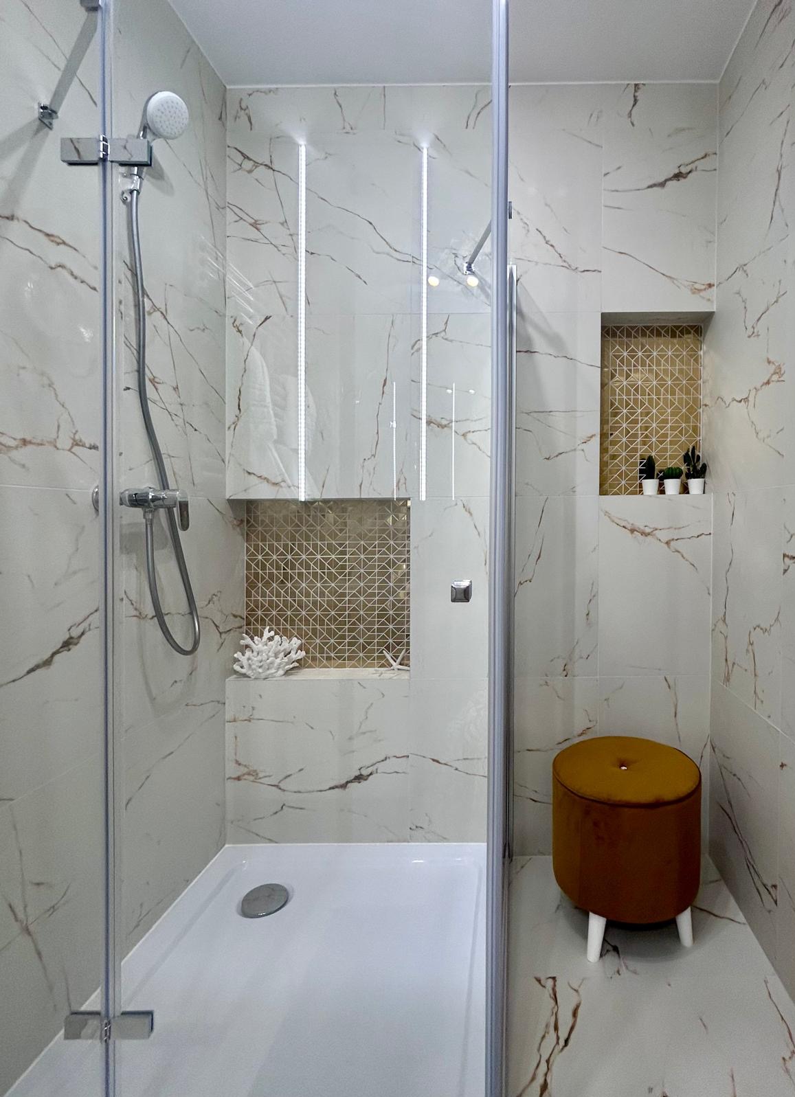 Nowoczesna kabina prysznicowa z białymi płytkami imitującymi marmur, złotymi akcentami w niszach, dekoracyjnym koralem i małym taboretem z aksamitu w kolorze musztardowym.