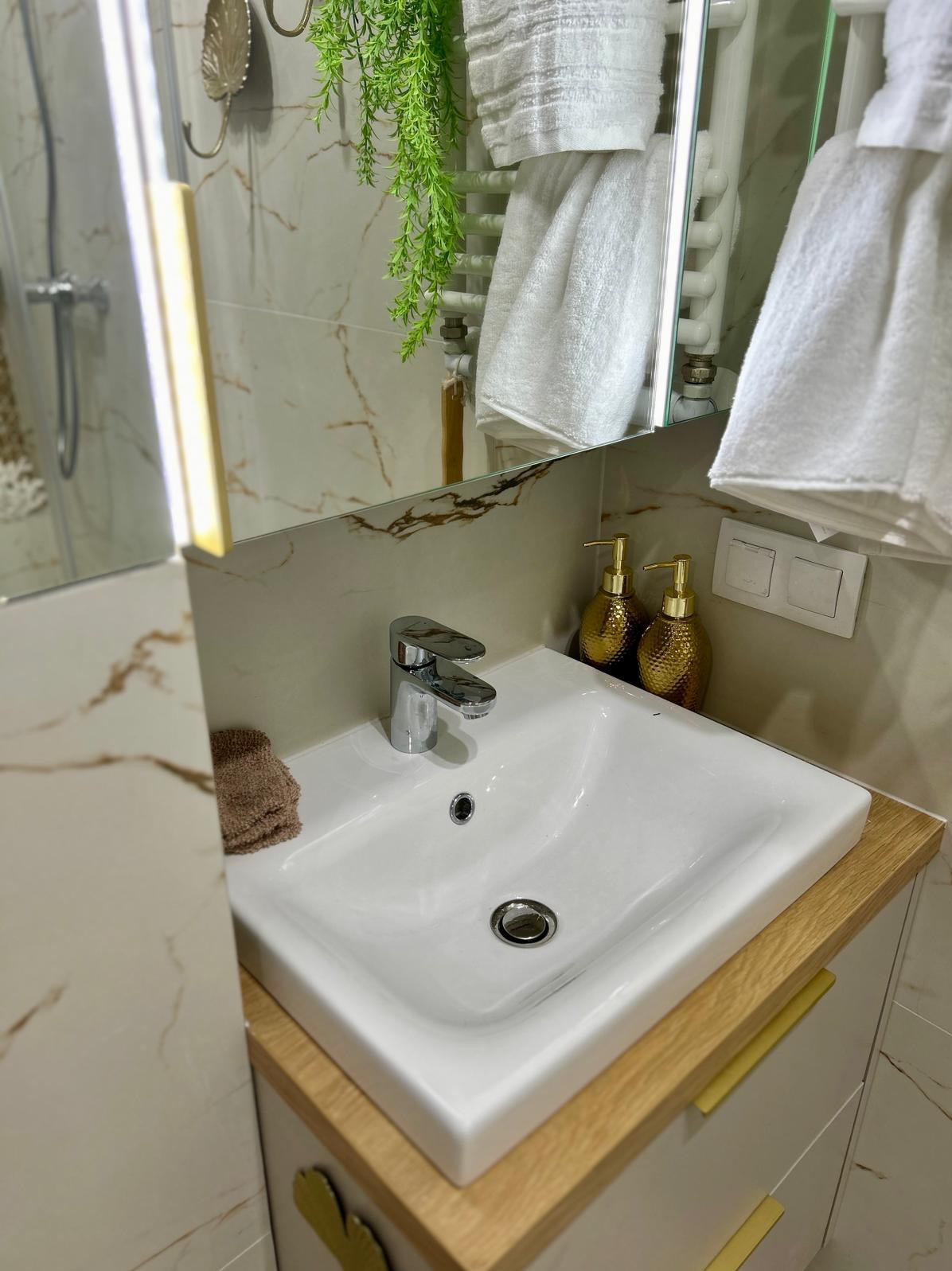 Biała umywalka nablatowa z chromowaną baterią, złotymi dozownikami do mydła i drewnianym blatem w łazience wykończonej marmurowymi płytkami.