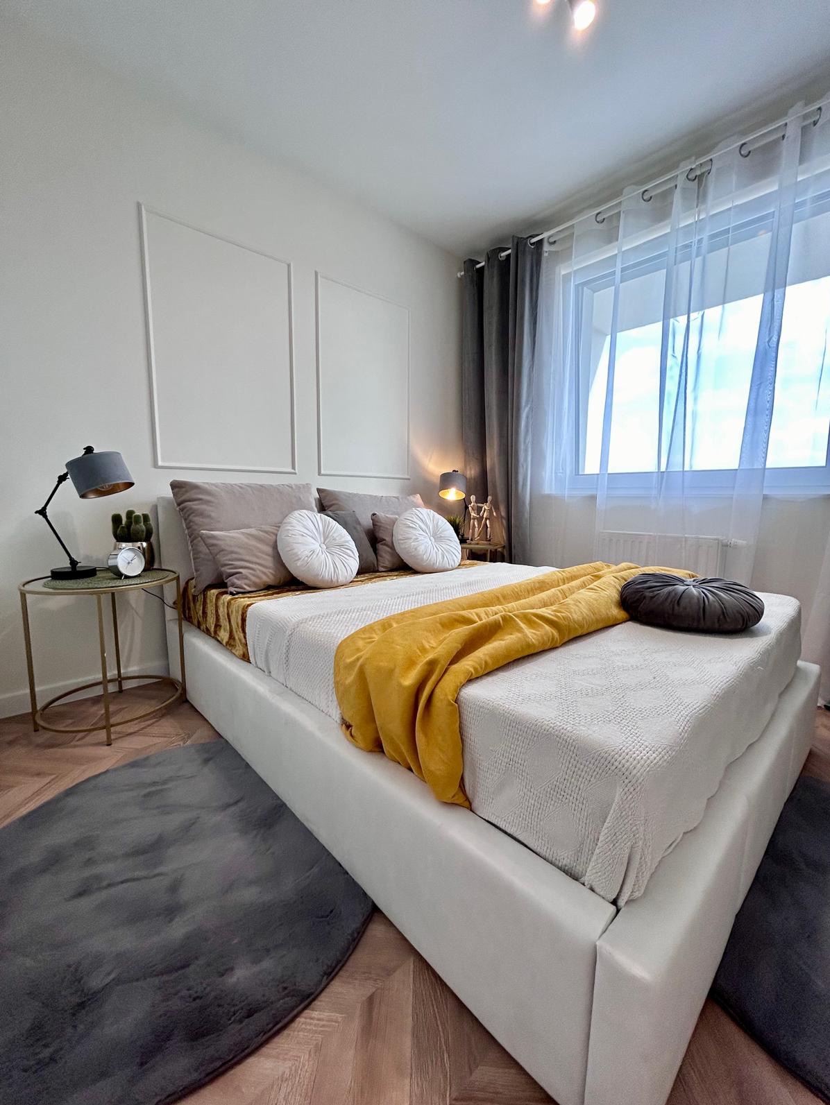 Nowoczesna sypialnia z białym łóżkiem kontynentalnym, żółtym pledem i okrągłymi poduszkami, uzupełniona o minimalistyczne stoliki nocne z lampkami i dekoracjami, na parkiecie w jodełkę i dywanie...