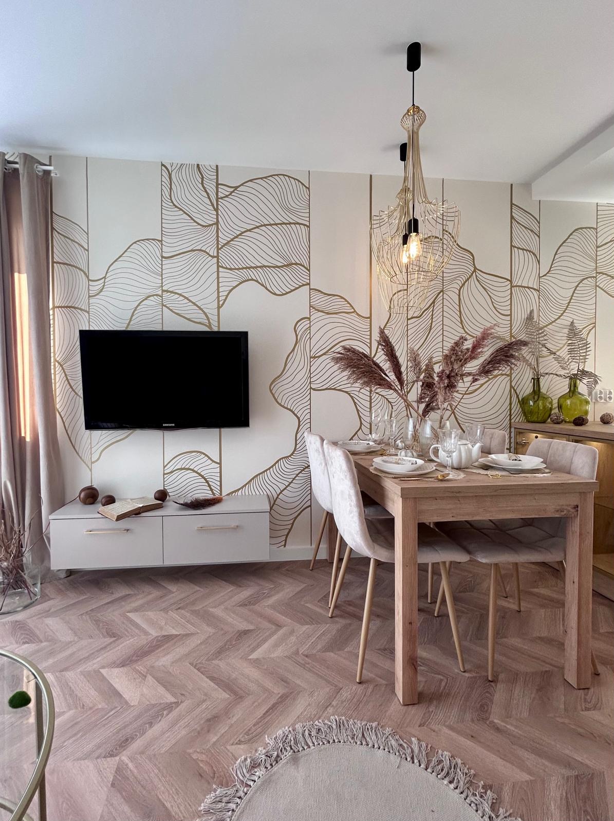 Jasne wnętrze salonu z jadalnią: drewniany stół nakryty do posiłku, welurowe krzesła, ściana z geometrycznym wzorem, wisząca lampa z metalowej siatki, telewizor na ścianie, parkiet w jodełkę.