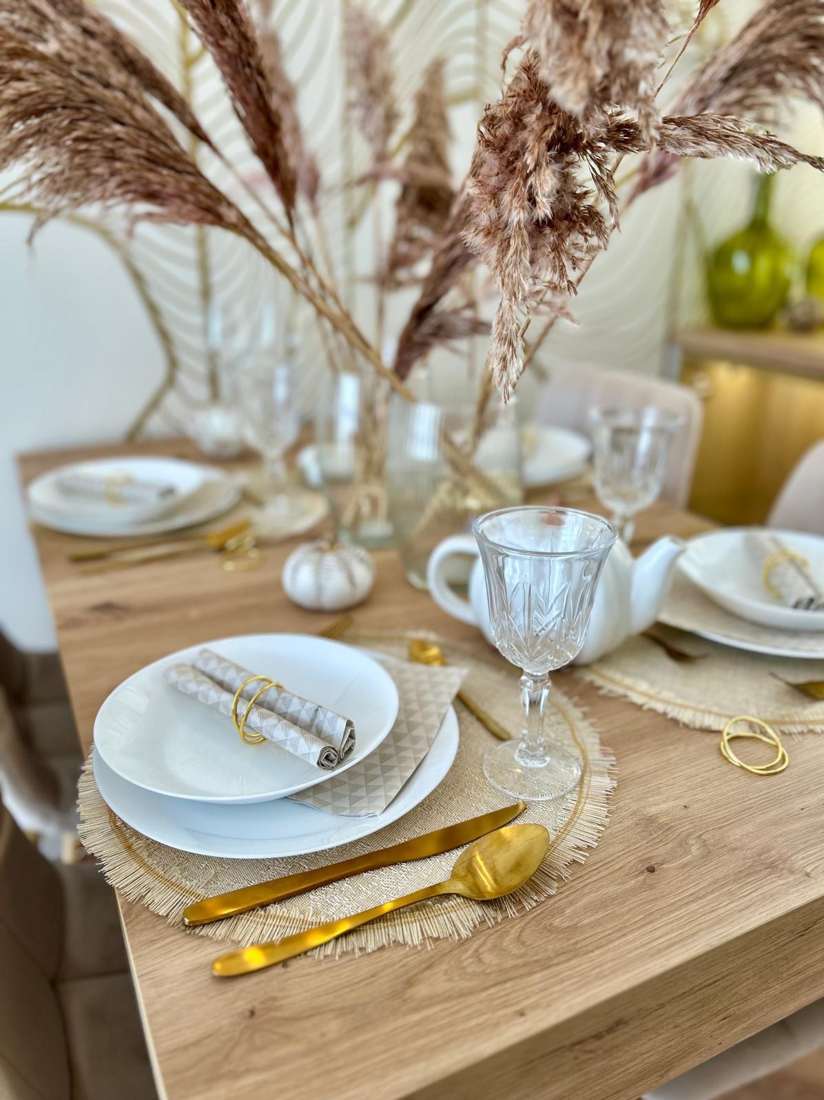 Uroczysty stół w Koluszkach, nakryty białymi talerzami z serwetkami w złotych pierścieniach, złotymi sztućcami, szklankami na nóżce oraz dekoracją z suszonych traw w wazonie, tworząc elegancką...