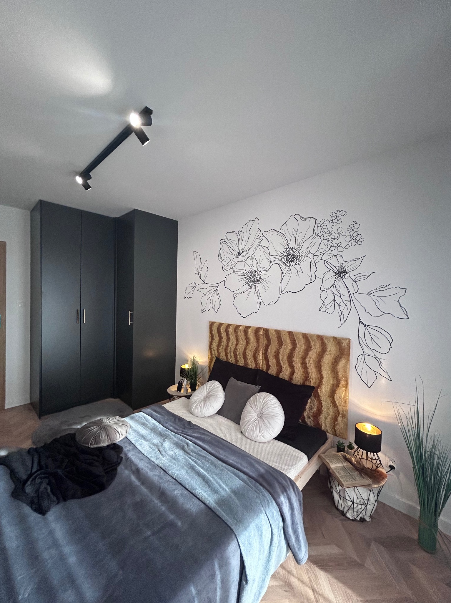 Nowoczesna sypialnia z ciemnymi meblami, drewnianym parkietem w jodełkę, łóżkiem z wezgłowiem w kolorze miodowym i kwiatowym motywem graficznym na ścianie.