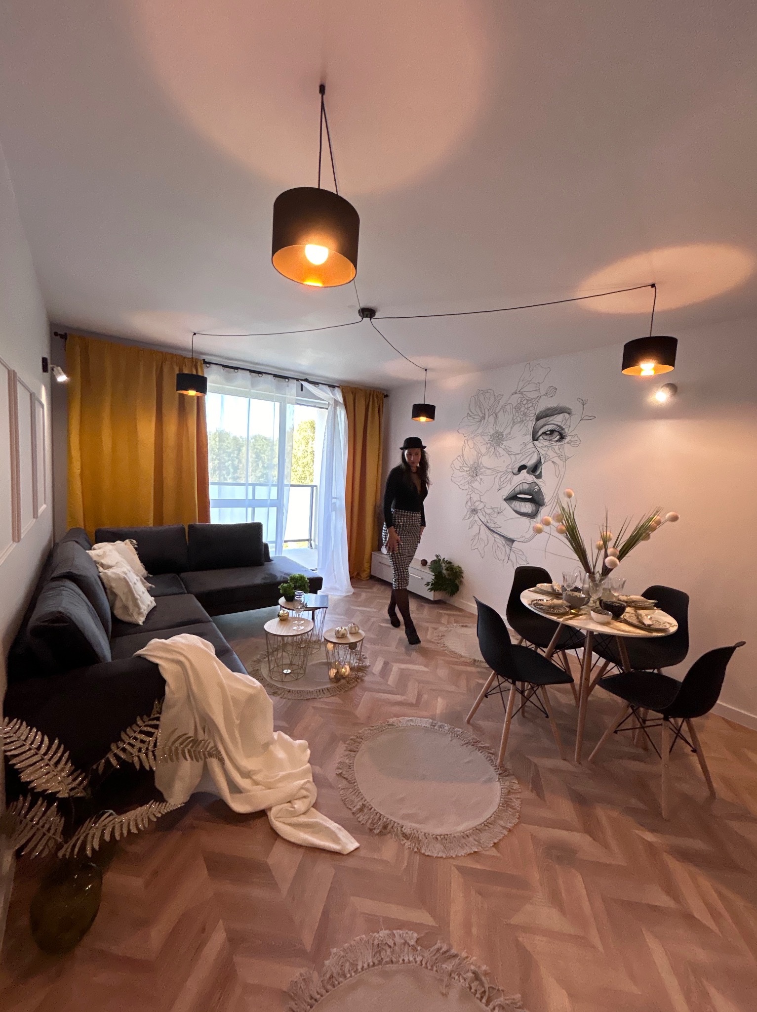 Nowoczesny salon w Koluszkach z szarym narożnikiem, okrągłym stołem z czarnymi krzesłami i muralem na ścianie. Kobieta w czarno-białym stroju i kapeluszu dodaje wnętrzu charakteru.