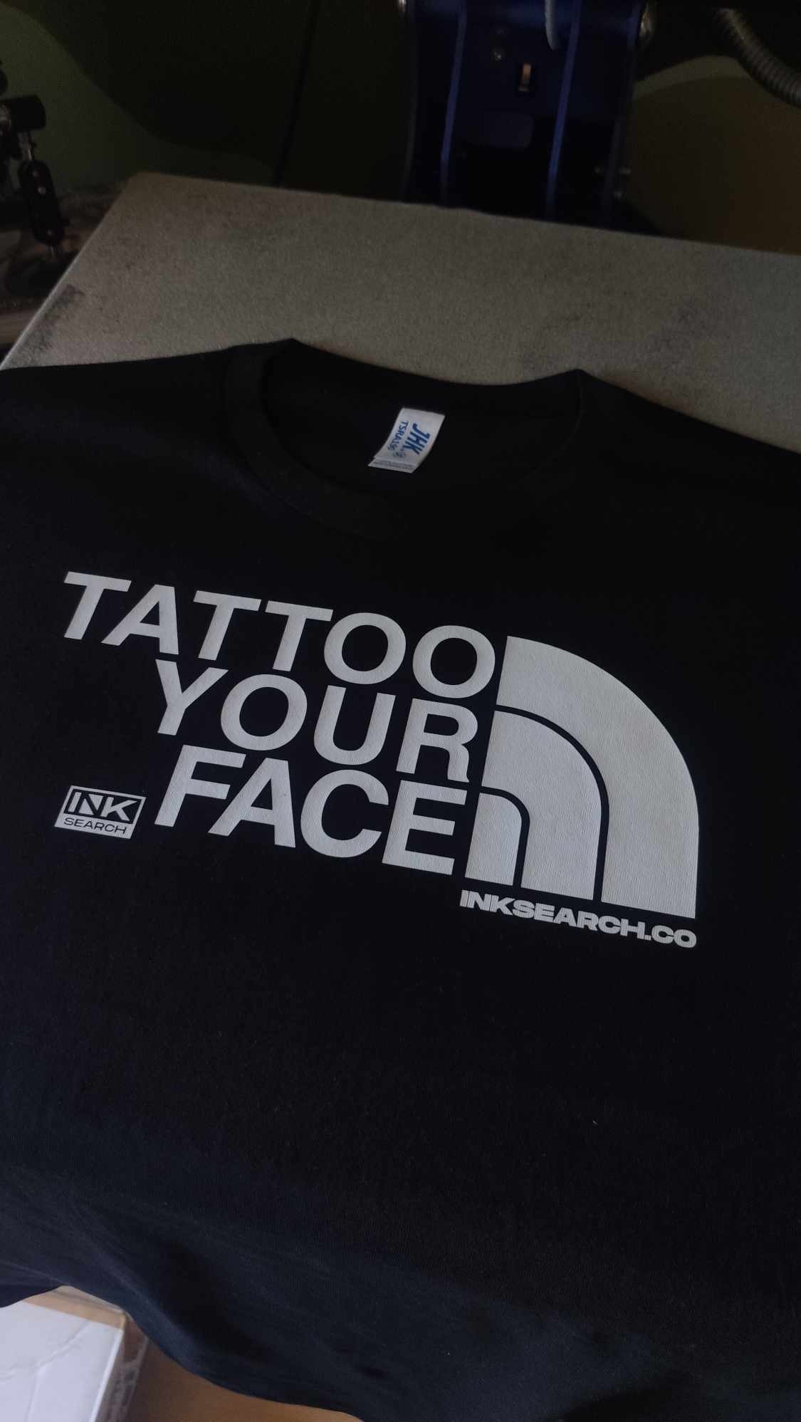 Czarna koszulka z białym nadrukiem 'TATTOO YOUR FACE' w stylu logo popularnej marki outdoorowej, obok logo 'INK SEARCH' i adresem strony internetowej.