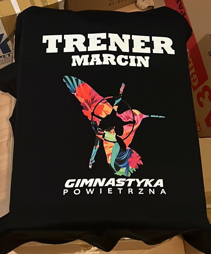 Czarna koszulka z nadrukiem: 'TRENER MARCIN', sylwetka gimnastyczki powietrznej wypełniona motywem kwiatowym, napis 'GIMNASTYKA POWIETRZNA'.