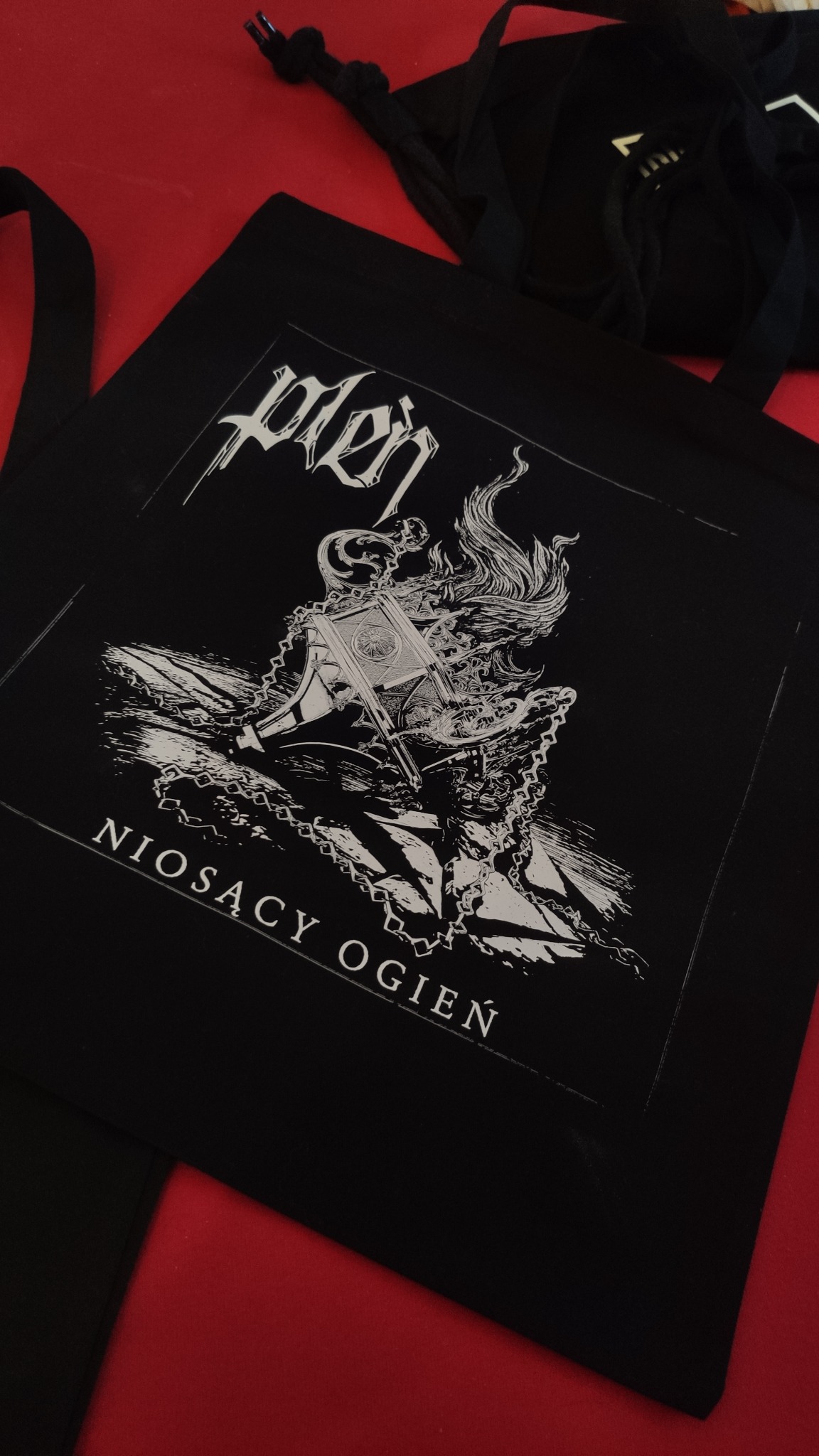 Czarna torba bawełniana z białym nadrukiem graficznym przedstawiającym stylizowaną postać i napis 'Niosący Ogień' na czerwonym tle.