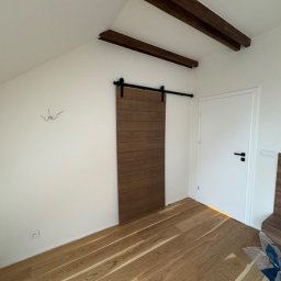 Deniro Denys Bykov - Drewniana, przesuwna brama w stylu stodoły na czarnym metalowym torze zamontowana w białym pokoju ze skośnym sufitem i drewnianymi belkami, jasna podłoga z parkietu.