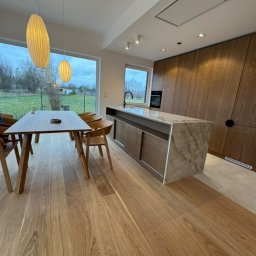 Deniro Denys Bykov - Jasna, przestronna jadalnia z widokiem na ogród, drewnianym parkietem, wyspą kuchenną z blatem imitującym marmur i wiszącymi lampami w stylu skandynawskim.