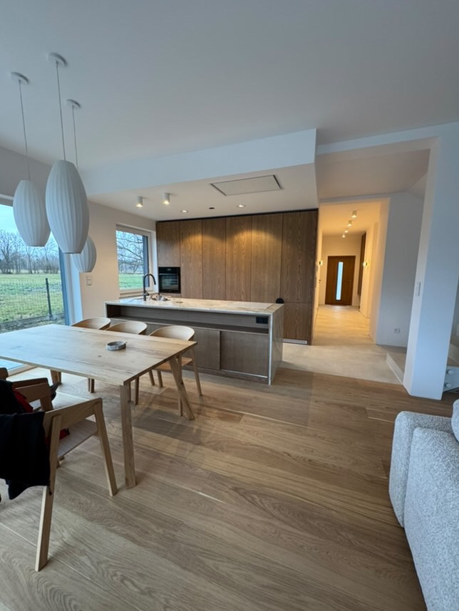 Jasne, przestronne wnętrze domu z drewnianą podłogą, stołem i krzesłami w jadalni, wyspą kuchenną z drewnianymi szafkami oraz widokiem na zewnątrz przez okno.