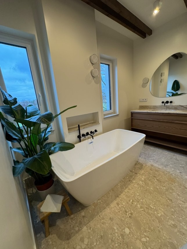 Nowoczesna łazienka z wolnostojącą białą wanną, armaturą w kolorze czarnym, lustrem o nieregularnym kształcie, drewnianą szafką pod umywalkę oraz podłogą z jasnego lastryko. Obok wanny stoi mały...