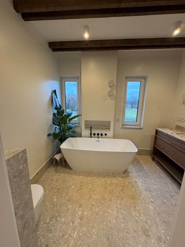 Nowoczesna łazienka z wolnostojącą białą wanną, jasnobeżową mozaikową podłogą i drewnianymi belkami stropowymi, oświetlona punktowymi lampami sufitowymi.