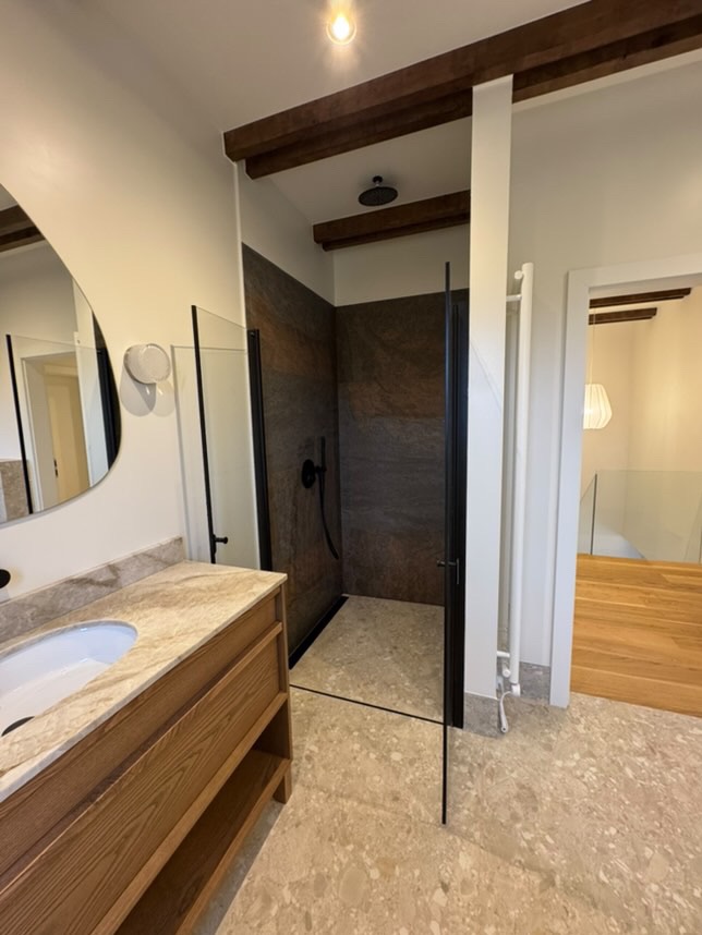 Nowoczesna łazienka z kabiną prysznicową typu walk-in, wykończona płytkami imitującymi kamień, z drewnianymi belkami stropowymi i umywalką z marmurowym blatem.