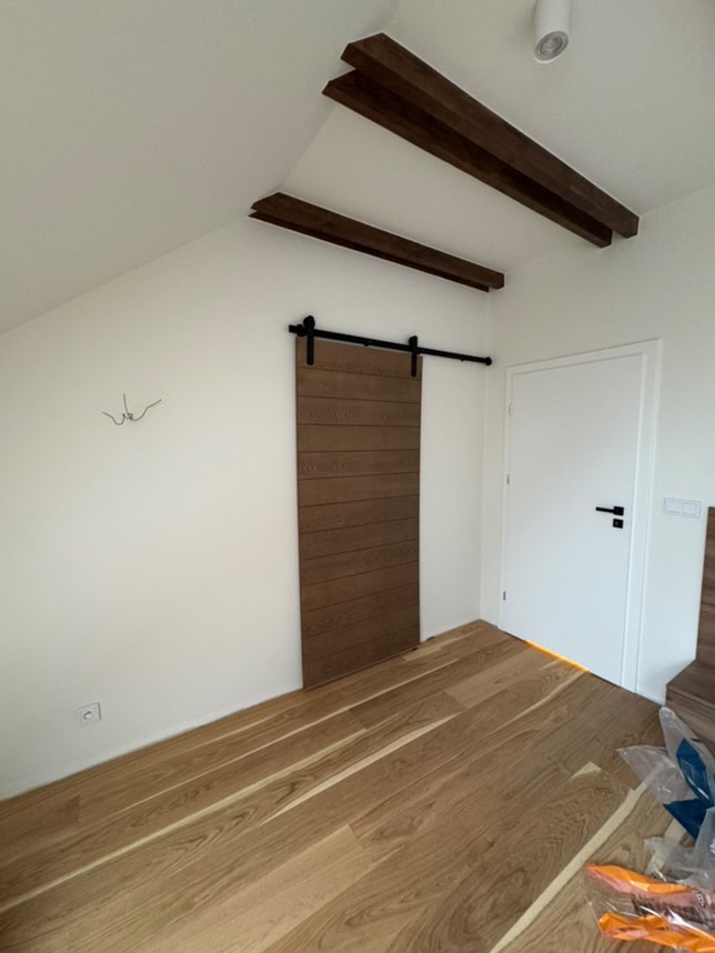 Drewniana, przesuwna brama w stylu stodoły na czarnym metalowym torze zamontowana w białym pokoju ze skośnym sufitem i drewnianymi belkami, jasna podłoga z parkietu.