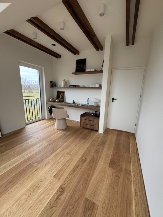 Jasny parkiet ułożony wzdłuż pokoju z drewnianymi belkami na suficie, widok na balkon i minimalistyczne meble.