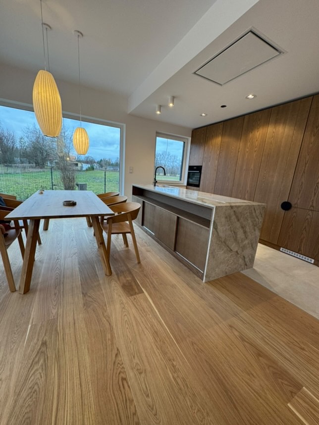 Jasna, przestronna jadalnia z widokiem na ogród, drewnianym parkietem, wyspą kuchenną z blatem imitującym marmur i wiszącymi lampami w stylu skandynawskim.