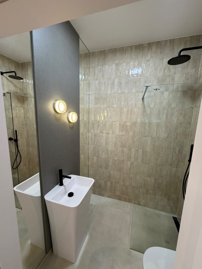 Nowoczesna łazienka z podłużnymi, beżowymi płytkami na ścianach, białą umywalką stojącą z czarną armaturą, deszczownicą i szklaną kabiną prysznicową.