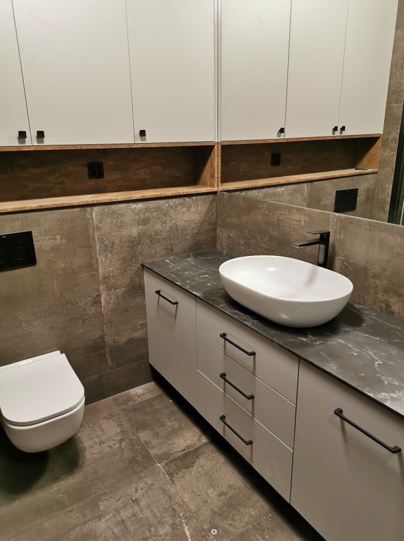 Nowoczesna łazienka z podwieszaną toaletą, szafką z umywalką nablatową i szarymi płytkami imitującymi kamień, uzupełniona szafkami wiszącymi z czarnymi uchwytami.